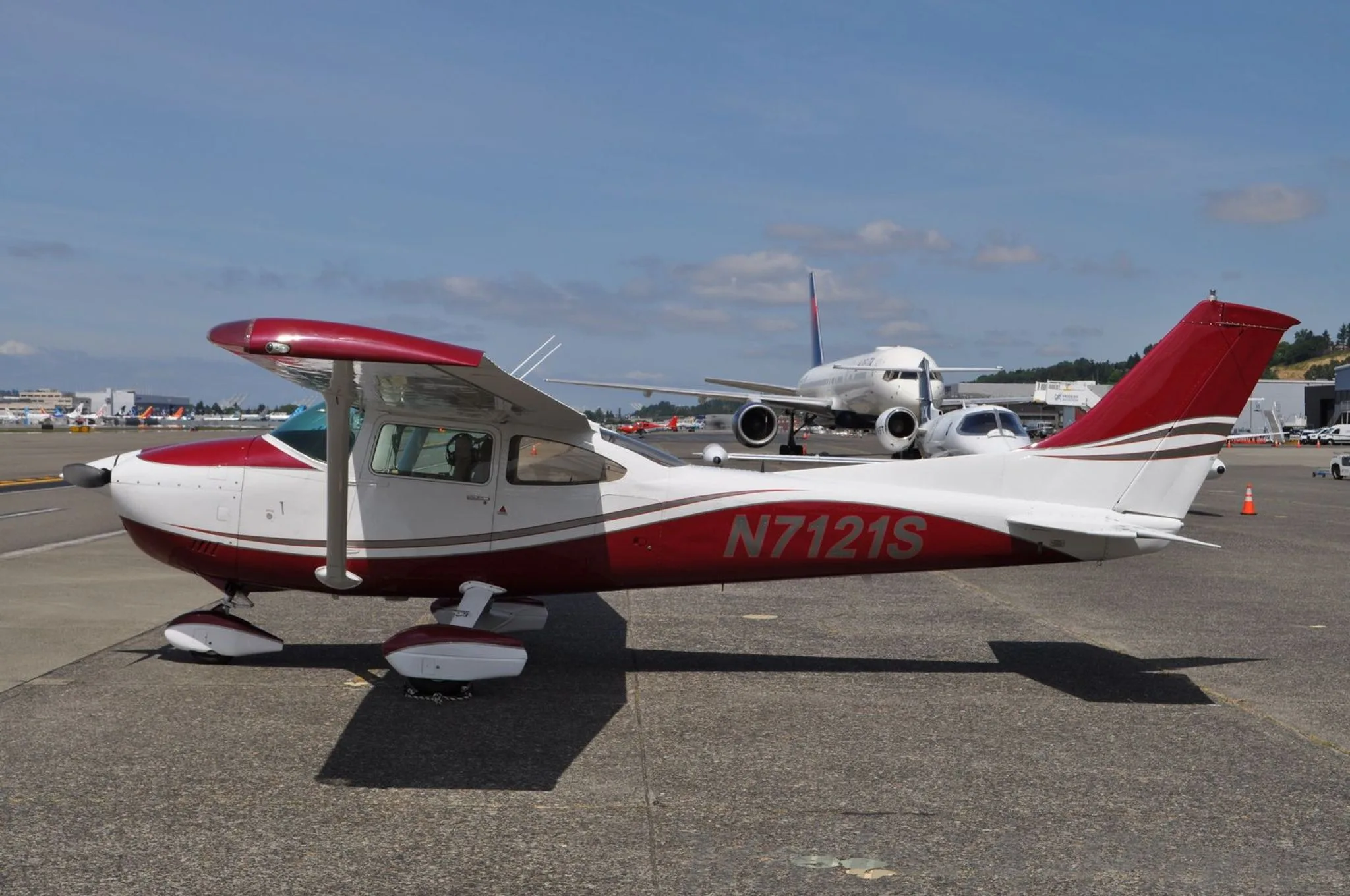 A red and white Cessna N7121S is parked on an airfield with a larger jet in the background.