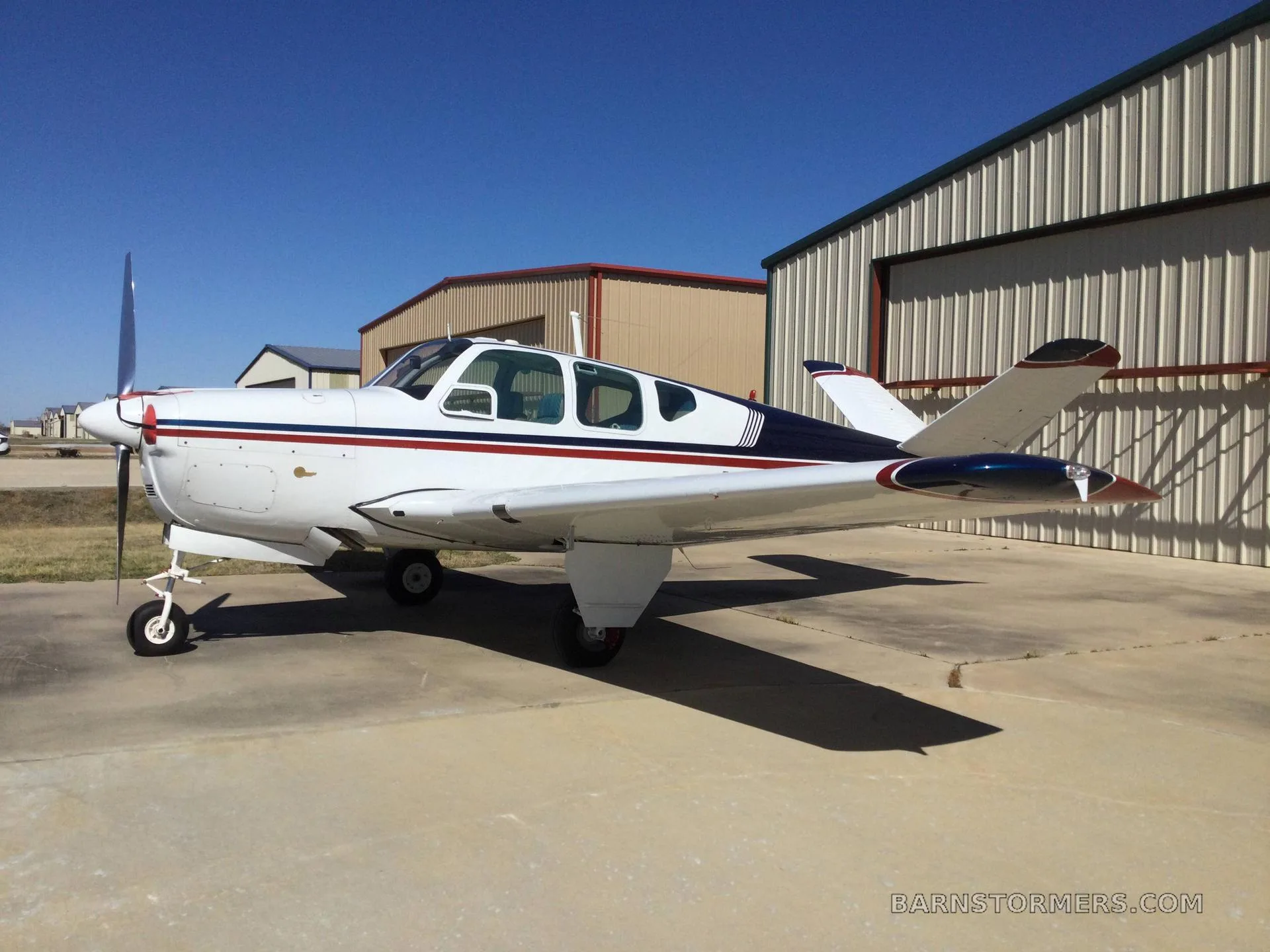A white aircraft with blue and red stripes is parked on a concrete pad next to hangars under a clear blue sky.