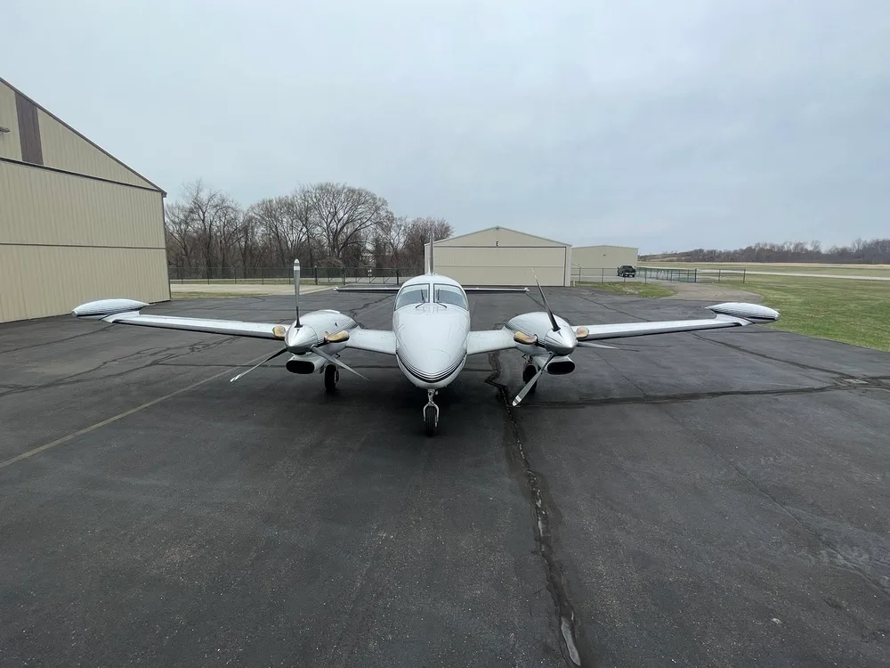 A front view of the white twin-engine aircraft N82TW with dark stripes, parked on a tarmac with hangars and a grassy field in the background.