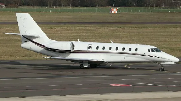 A white business jet with a dark stripe on its fuselage is seen from the side on an airport tarmac.