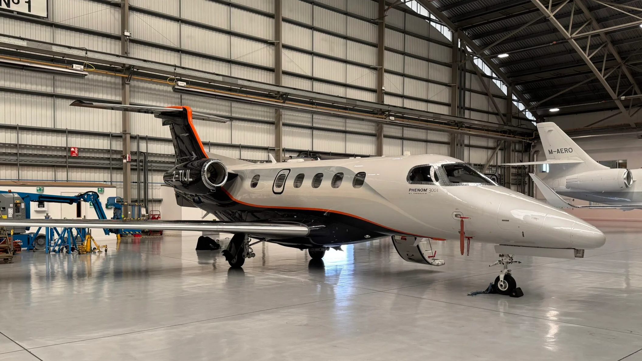 An Embraer Phenom 300E business jet with a white, black, and orange paint scheme is parked inside a hangar.