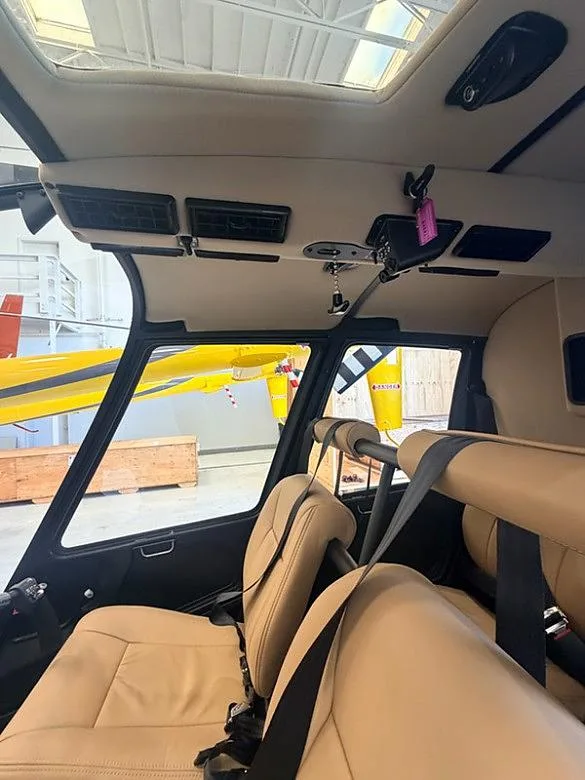 The tan leather interior cabin of a helicopter, looking forward with a yellow helicopter visible outside.