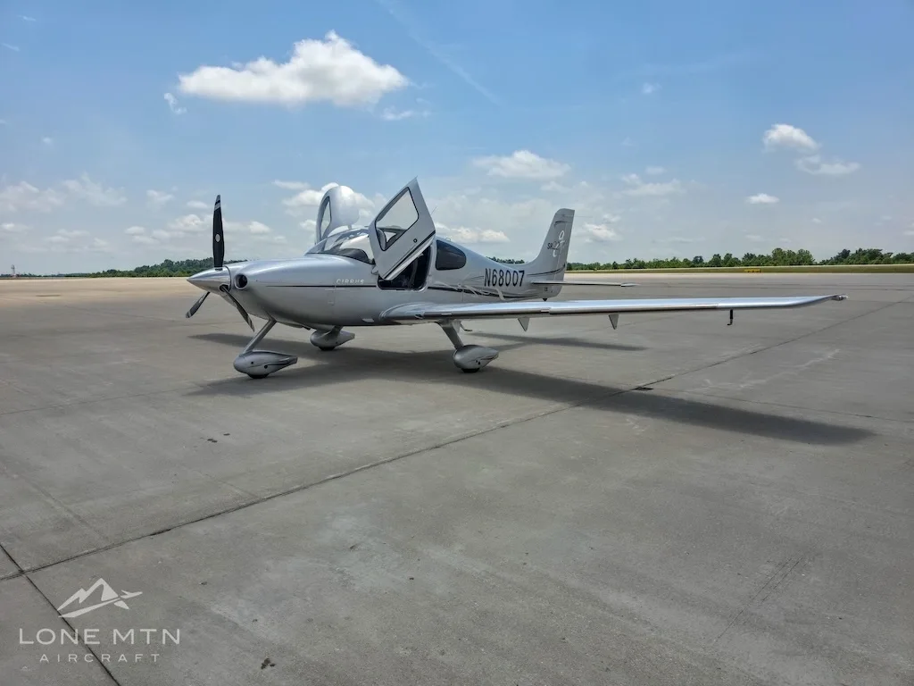 A silver Cirrus SR22 aircraft (N68007) with its gull-wing doors open, parked on an airfield under a partly cloudy sky.