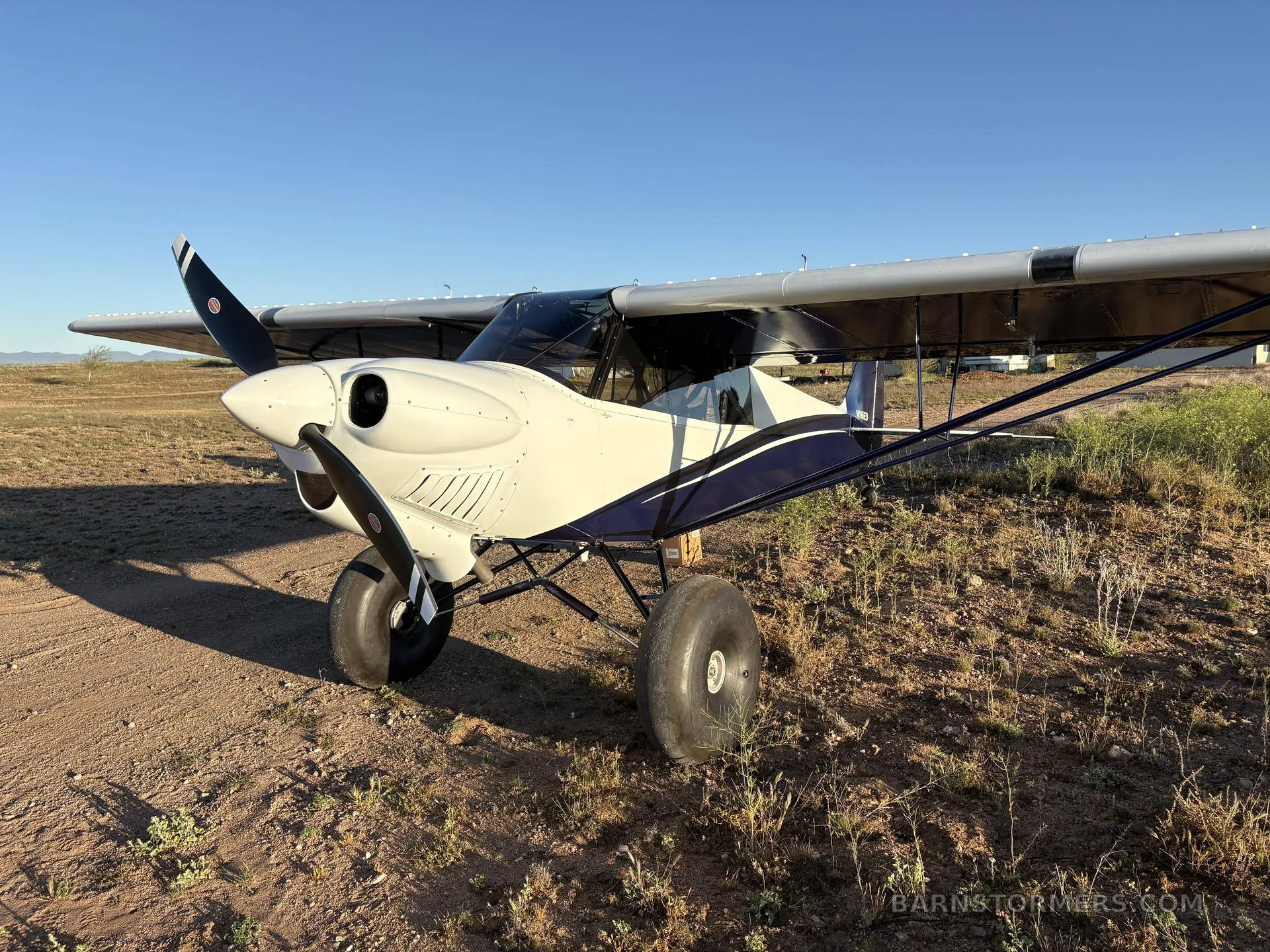 A white and blue CubCrafters aircraft with large tundra tires parked on a dirt field under a clear sky.