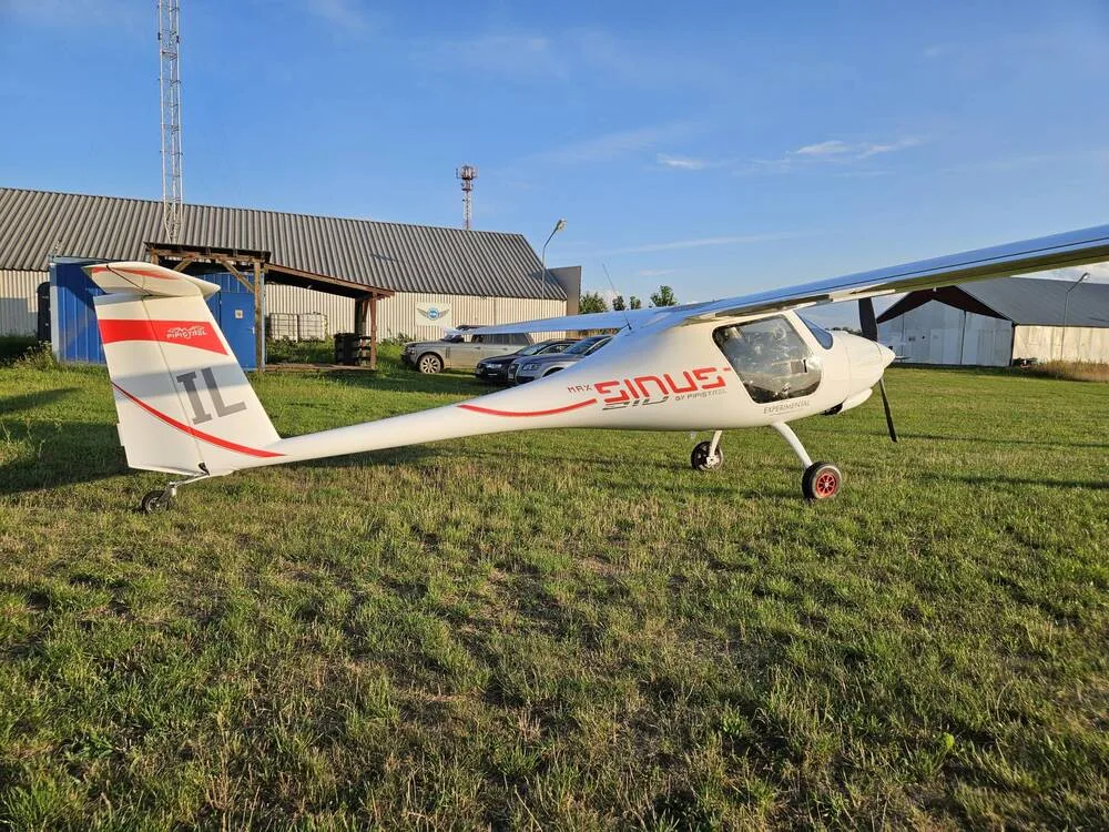 A white Pipistrel Sinus aircraft with red accents and "IL" tail number is parked on a grassy airfield next to hangars.