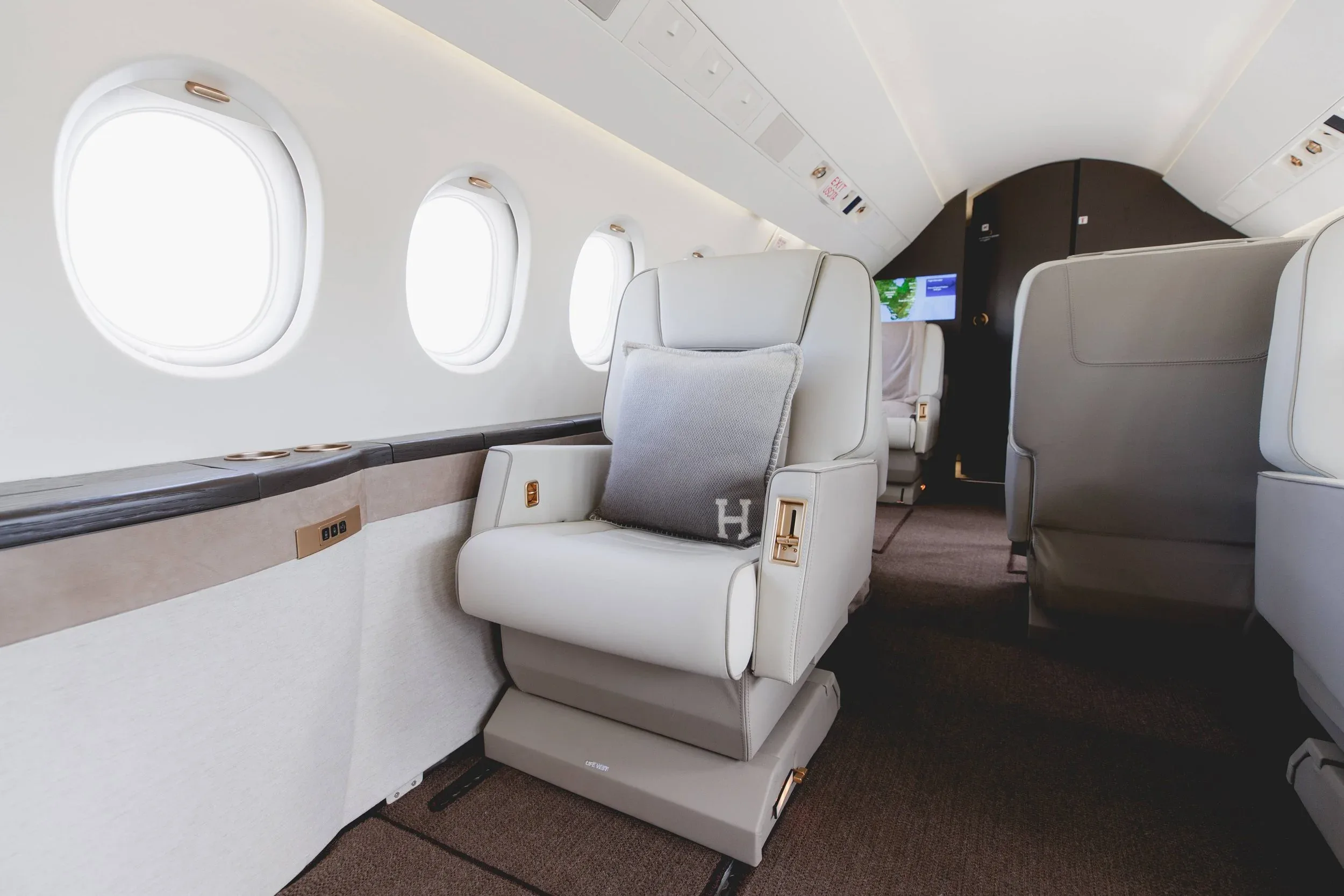 A close-up view of a plush light-colored leather seat with an "H" monogrammed pillow inside a private jet cabin.