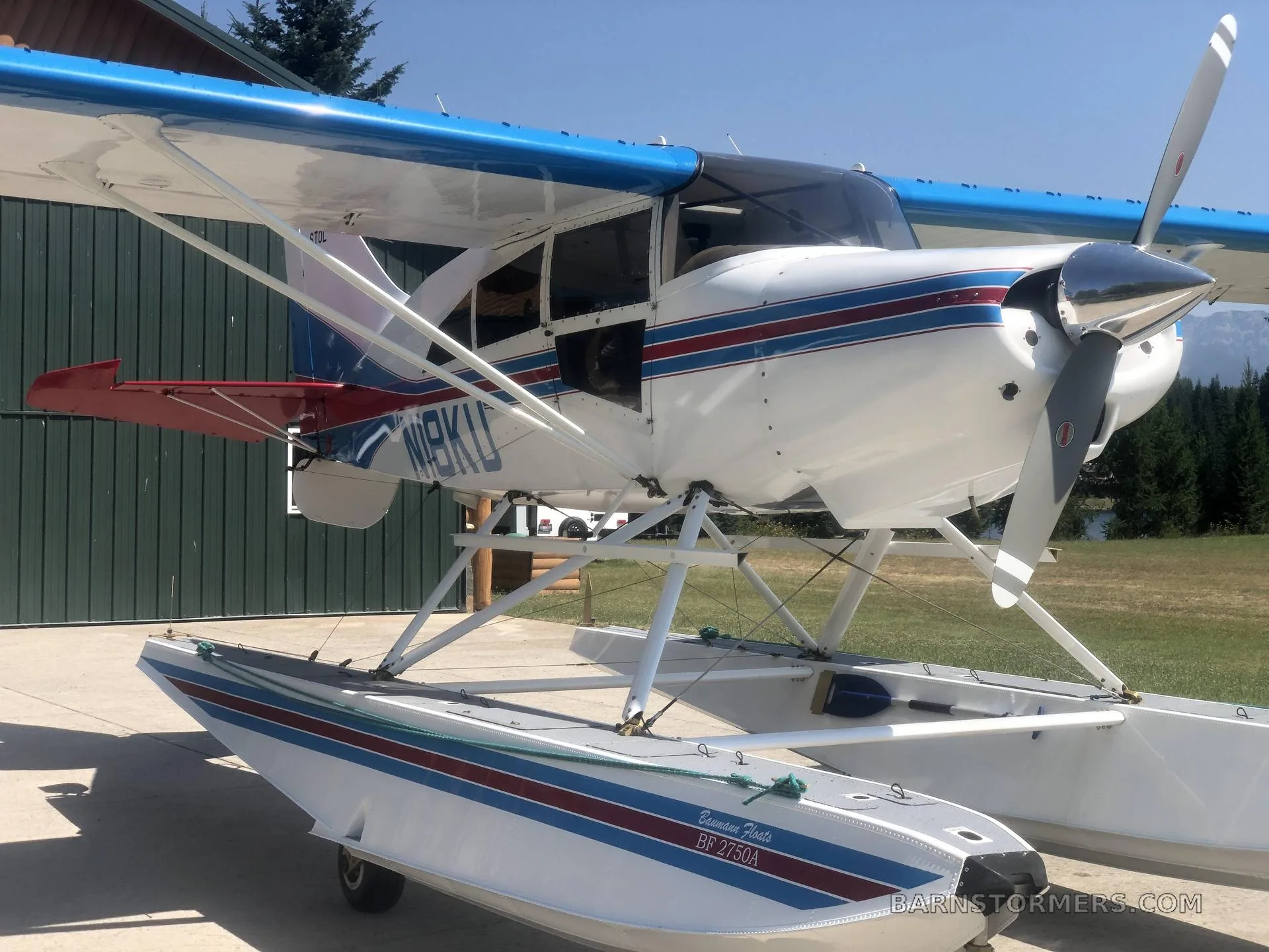 A white and blue floatplane, N18KU, with red tail accents, parked on concrete next to a green building.