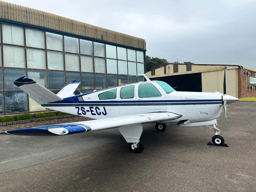 A white and blue Beechcraft Bonanza parked on an tarmac next to a hangar.