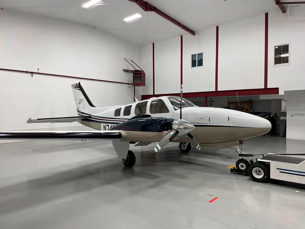 A white and tan twin-engine aircraft with dark blue accents is parked in a well-lit hangar.
