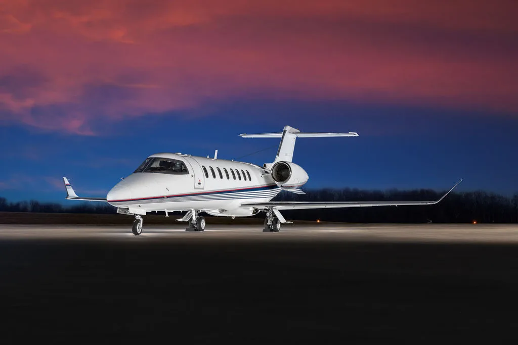 A white business jet with blue and red stripes on the fuselage is parked on an airfield at sunset.