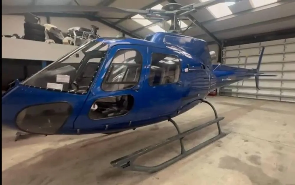 A blue helicopter, possibly an Airbus H125, with its main rotor blades removed, parked inside a hangar.