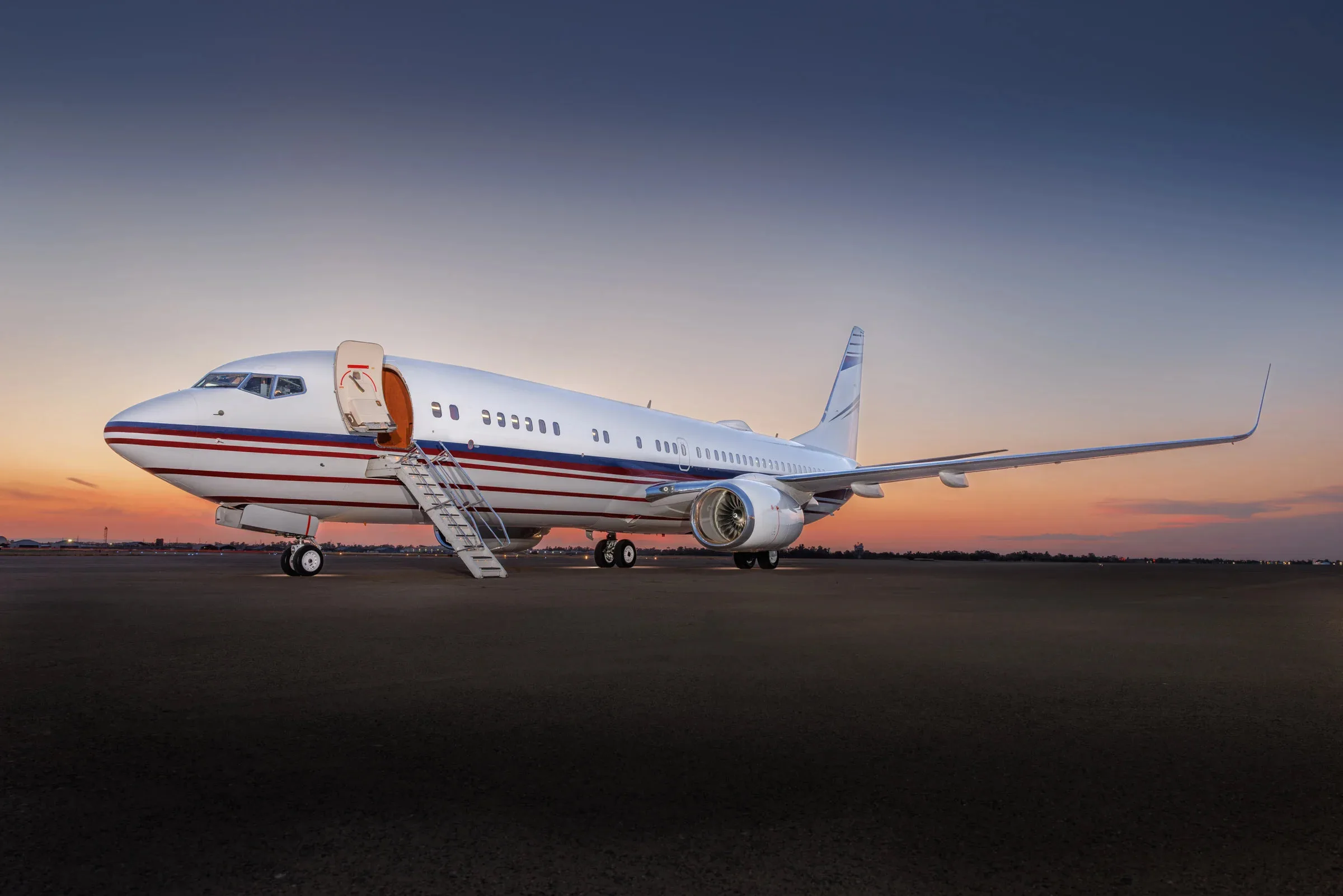 A white Boeing 737 business jet with red and blue stripes on the fuselage and tail, parked on an tarmac at sunset with its main door open.