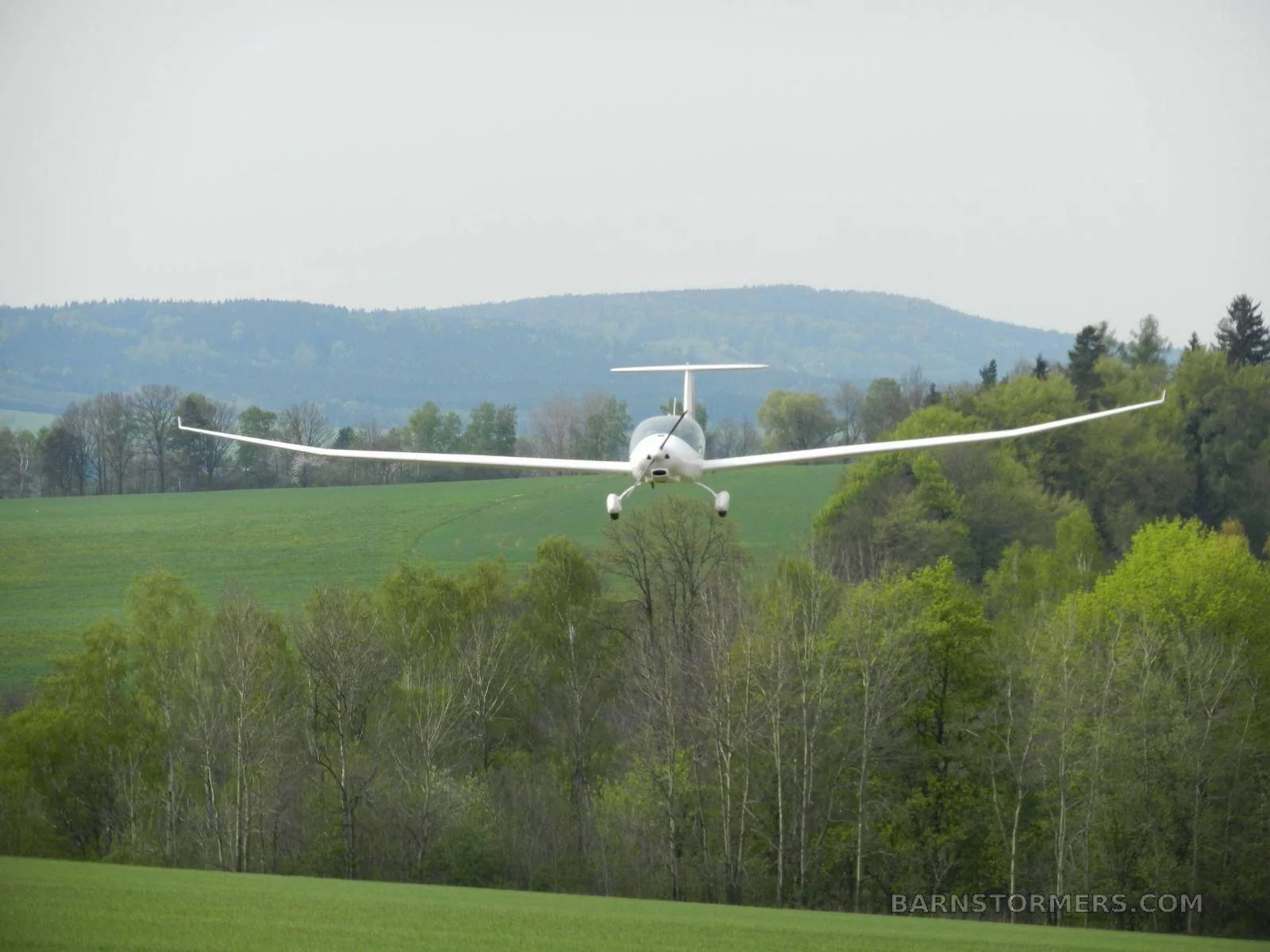 A white motor glider with landing gear down flies low over a green field with trees and hills.