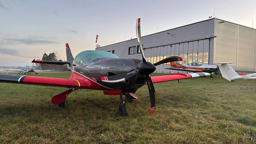 A grey and red BOSSA 915iS aircraft with a propeller, parked on a grassy field in front of a hangar.