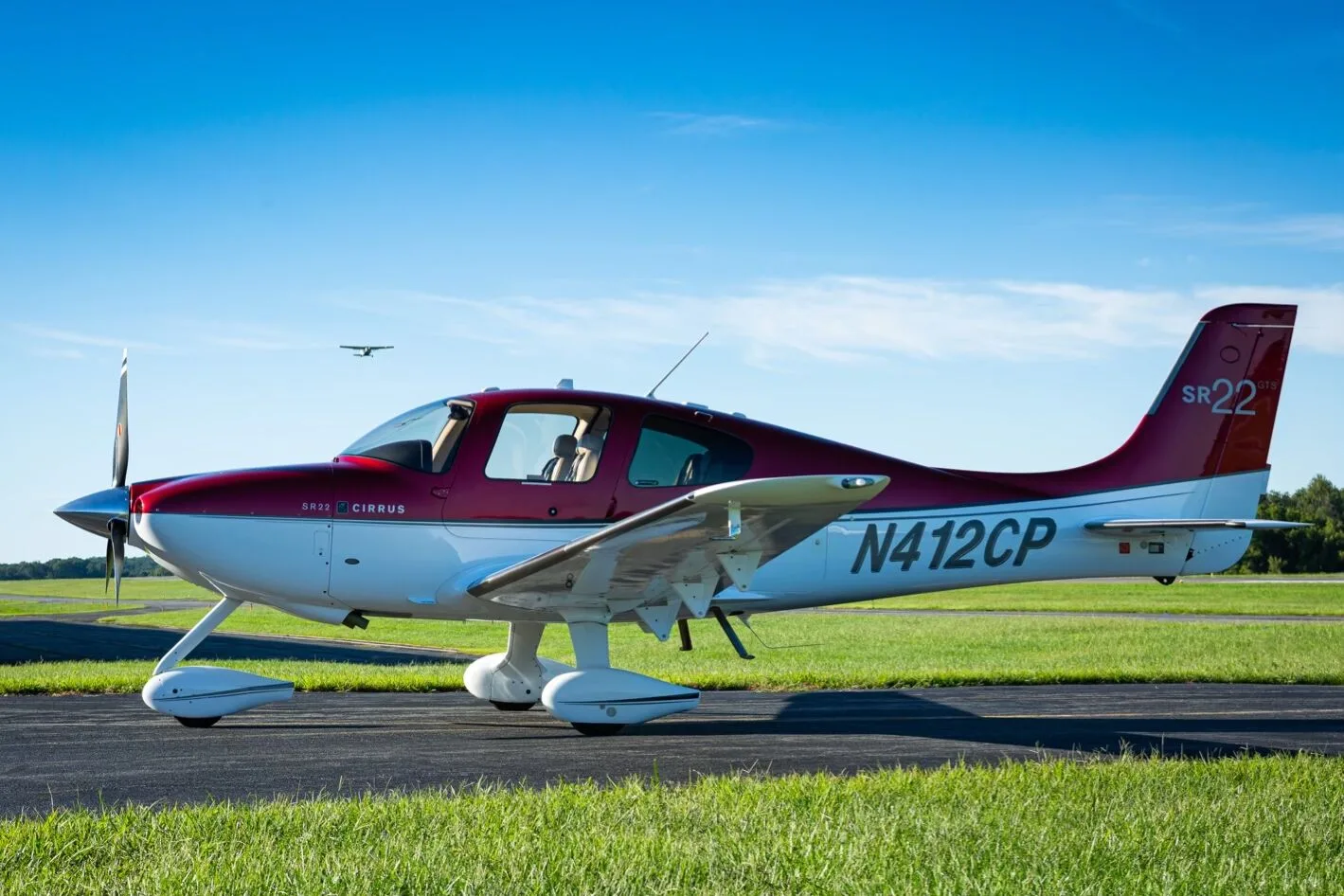 A red and white Cirrus SR22 aircraft (N412CP) parked on an airfield with another aircraft flying in the distance.