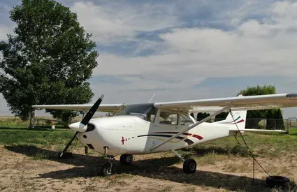 A white single-engine propeller aircraft with dark and red stripes is parked on a dirt field with a large tree in the background.