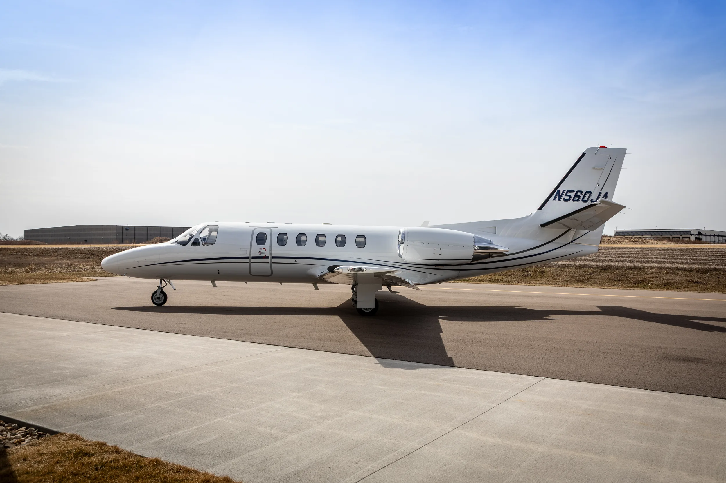 A white Cessna Citation Excel (N560JA) with black and grey stripes parked on an airfield under a partly cloudy sky.