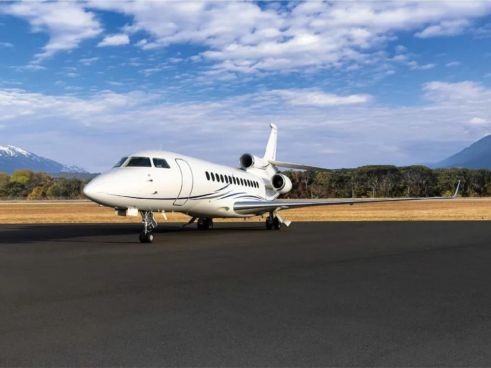 A white Dassault Falcon business jet with blue stripes parked on an airfield with mountains in the background.