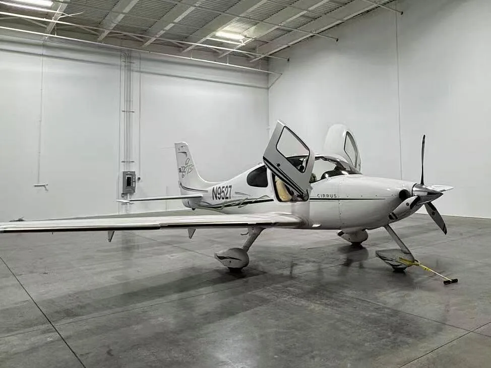 A white Cirrus aircraft with registration N9527 is parked in a hangar with its doors open.