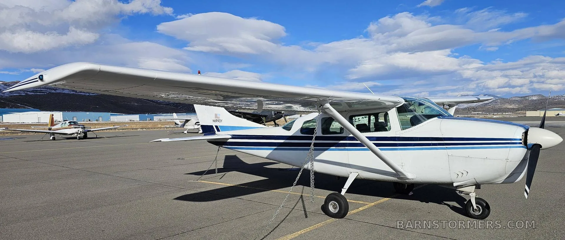 A white and blue Cessna 182 aircraft with registration N6990Y parked on an airfield, viewed from the side.
