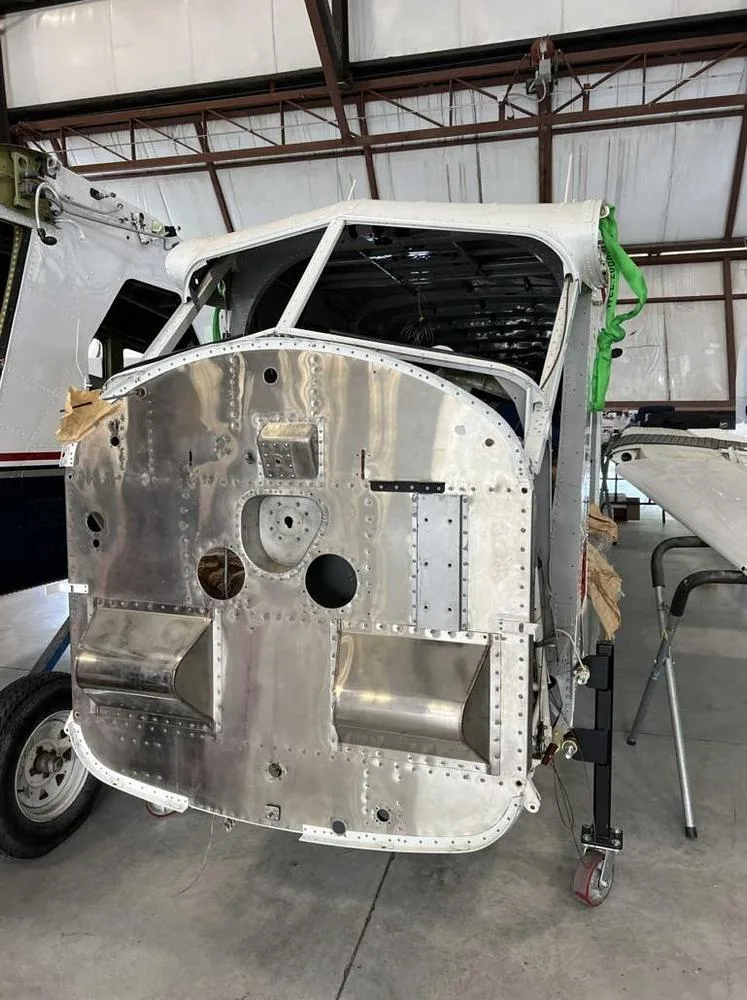 The unpainted metal nose section of an aircraft, showing the cockpit frame and riveted panels, in a hangar.