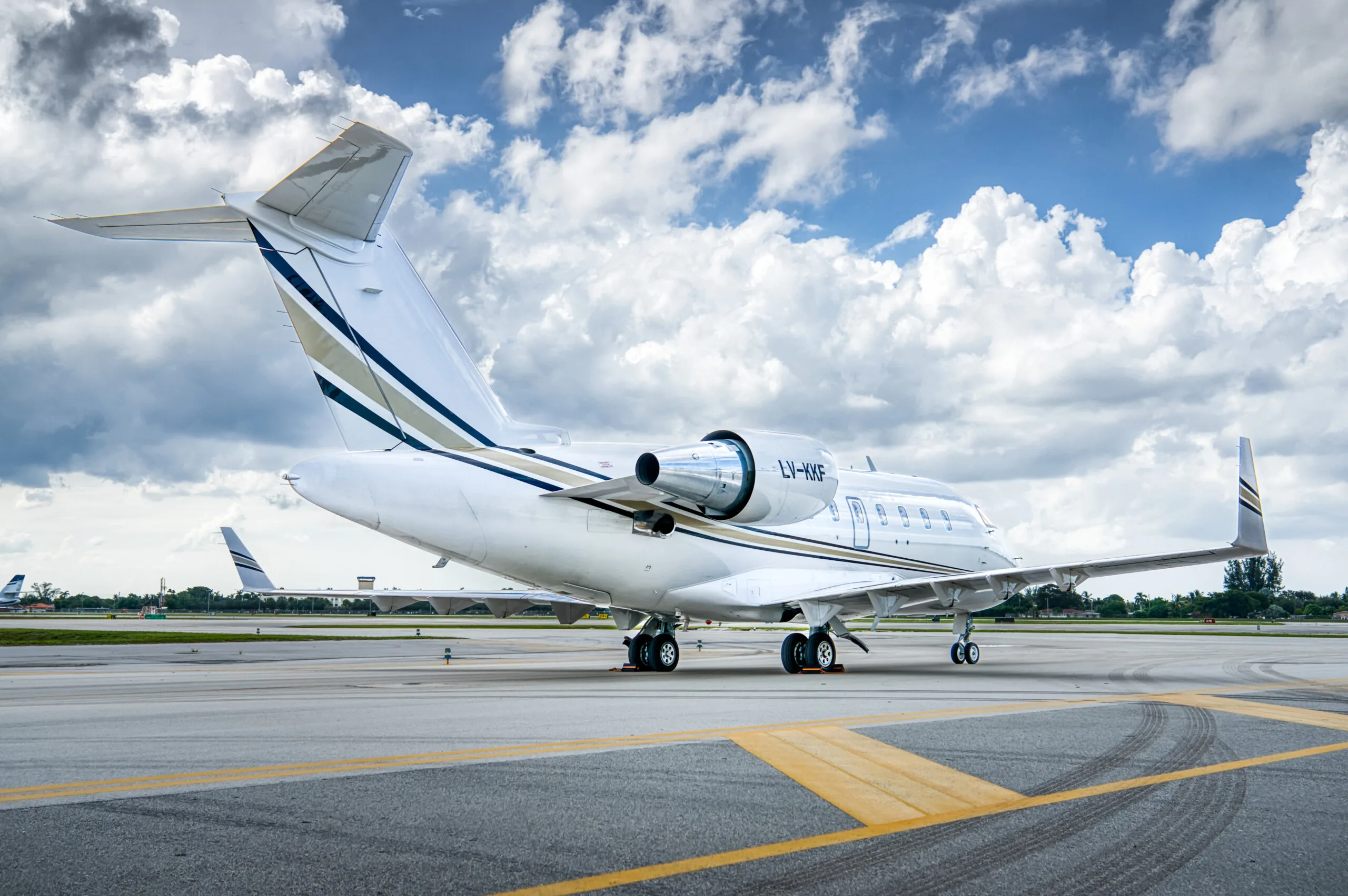 A white Bombardier Challenger business jet with gold and black stripes, registration LV-KKF, parked on an airport tarmac under a cloudy sky.