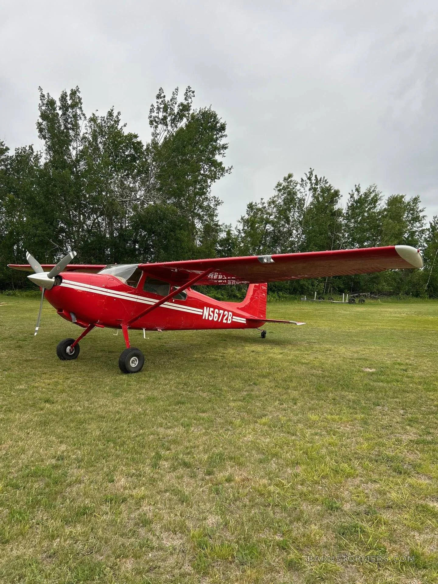 A side view of a red single-engine aircraft with tail number N5672B parked on a grassy field.