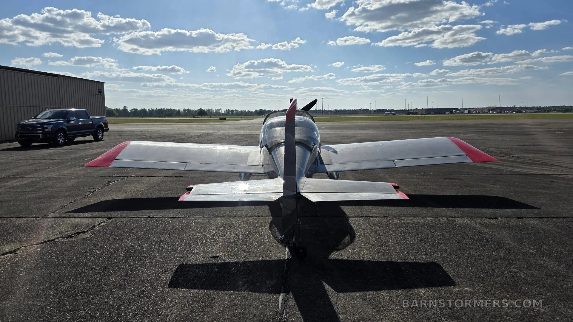 A rear view of a silver single-engine aircraft with red wingtips and tail accents, parked on an asphalt tarmac with a pickup truck and airport buildings in the distance.