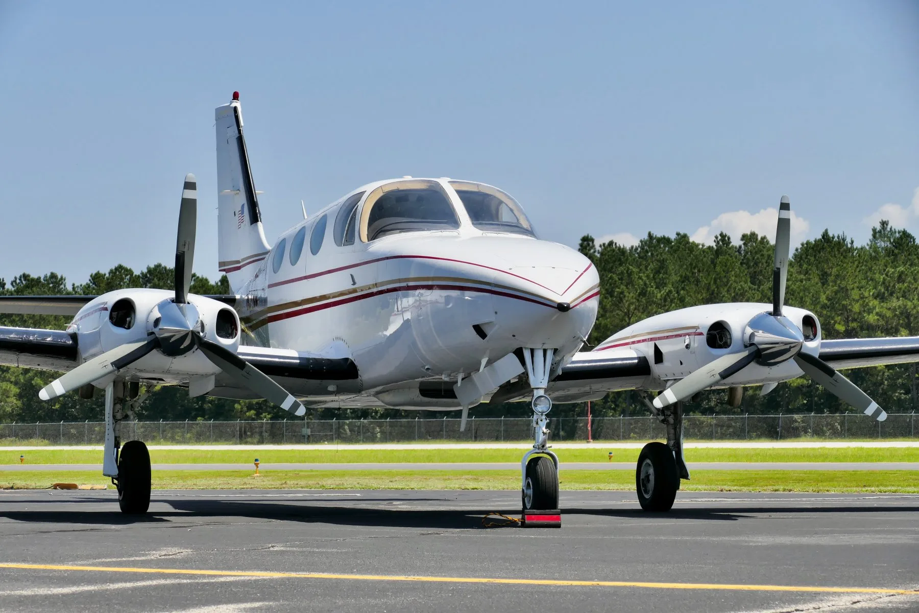 Front view of a white twin-engine propeller aircraft with red and gold stripes on an airfield.