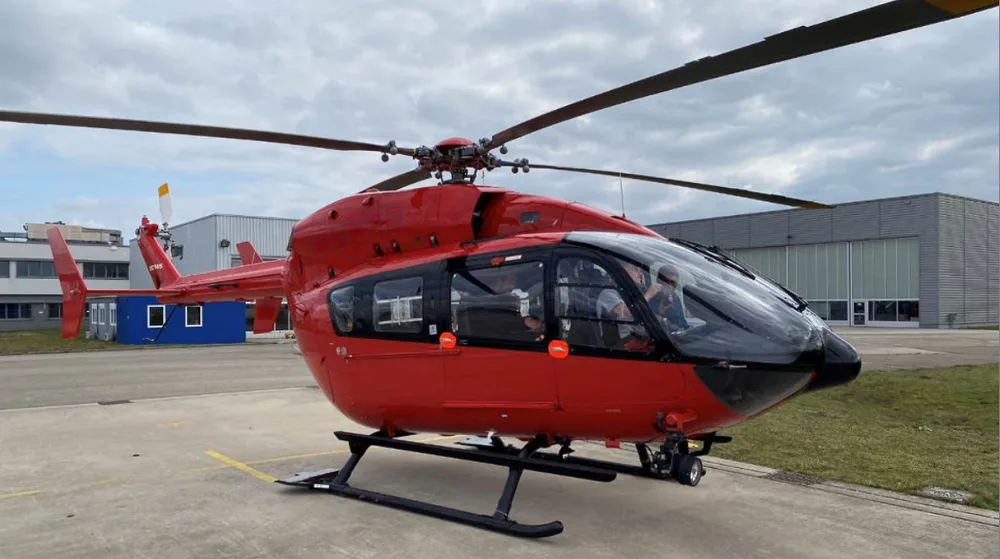 A red Airbus H145 helicopter is parked on an asphalt apron with hangars in the background.