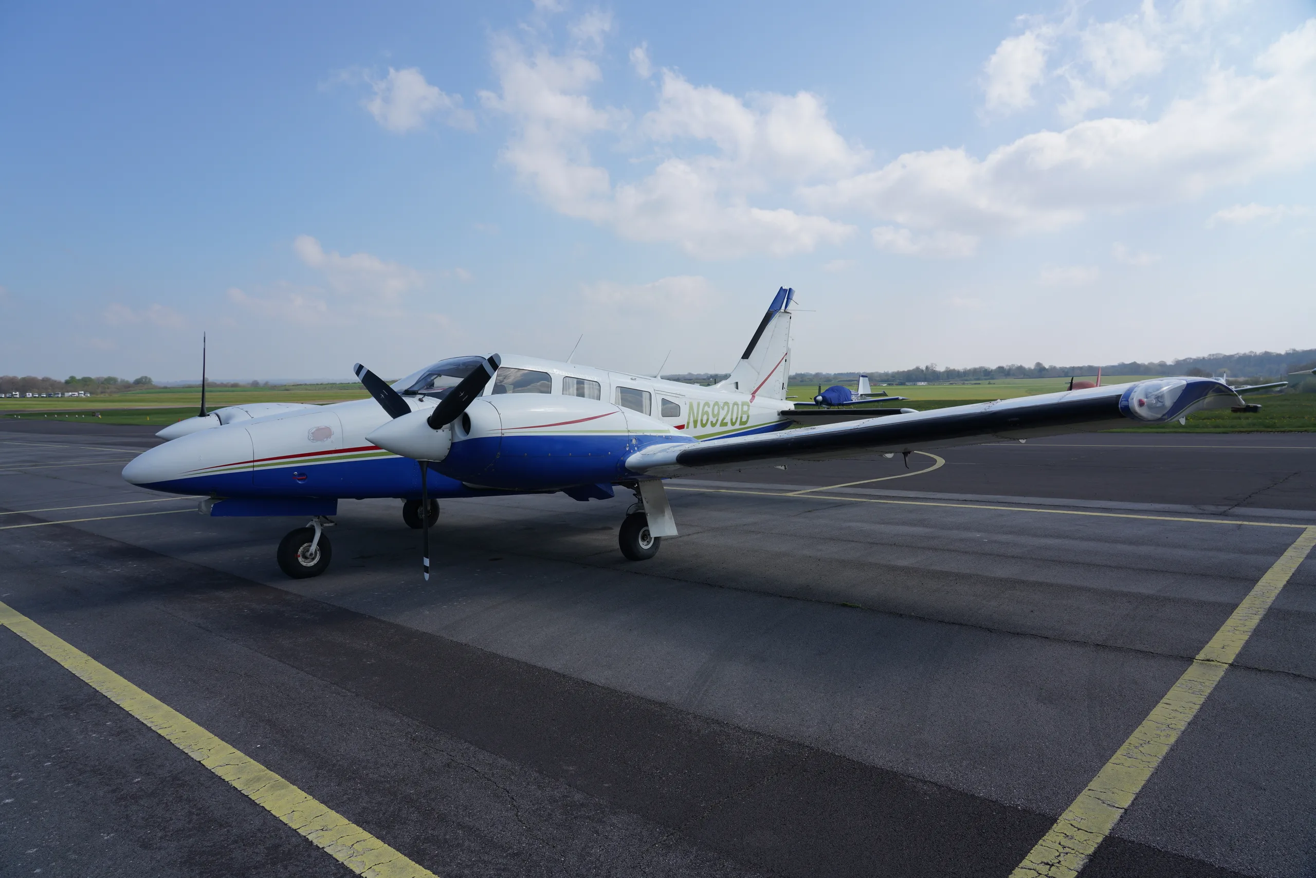 A white and blue twin-engine Piper Seneca aircraft with green and red stripes parked on an asphalt tarmac under a partly cloudy sky.