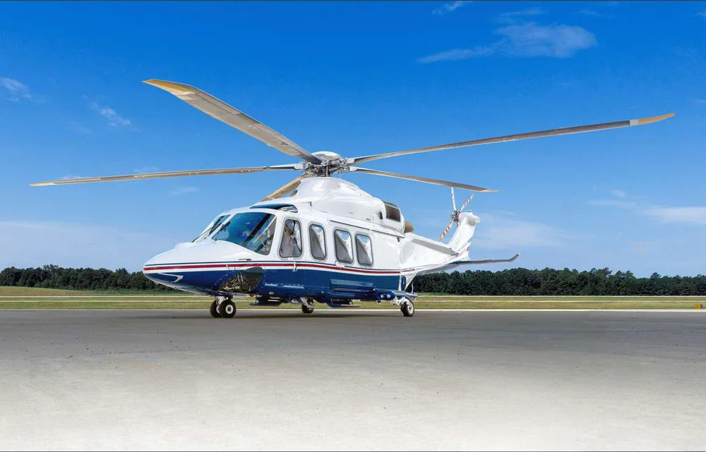 A white and blue helicopter with multiple rotor blades sits on an asphalt tarmac under a clear blue sky.