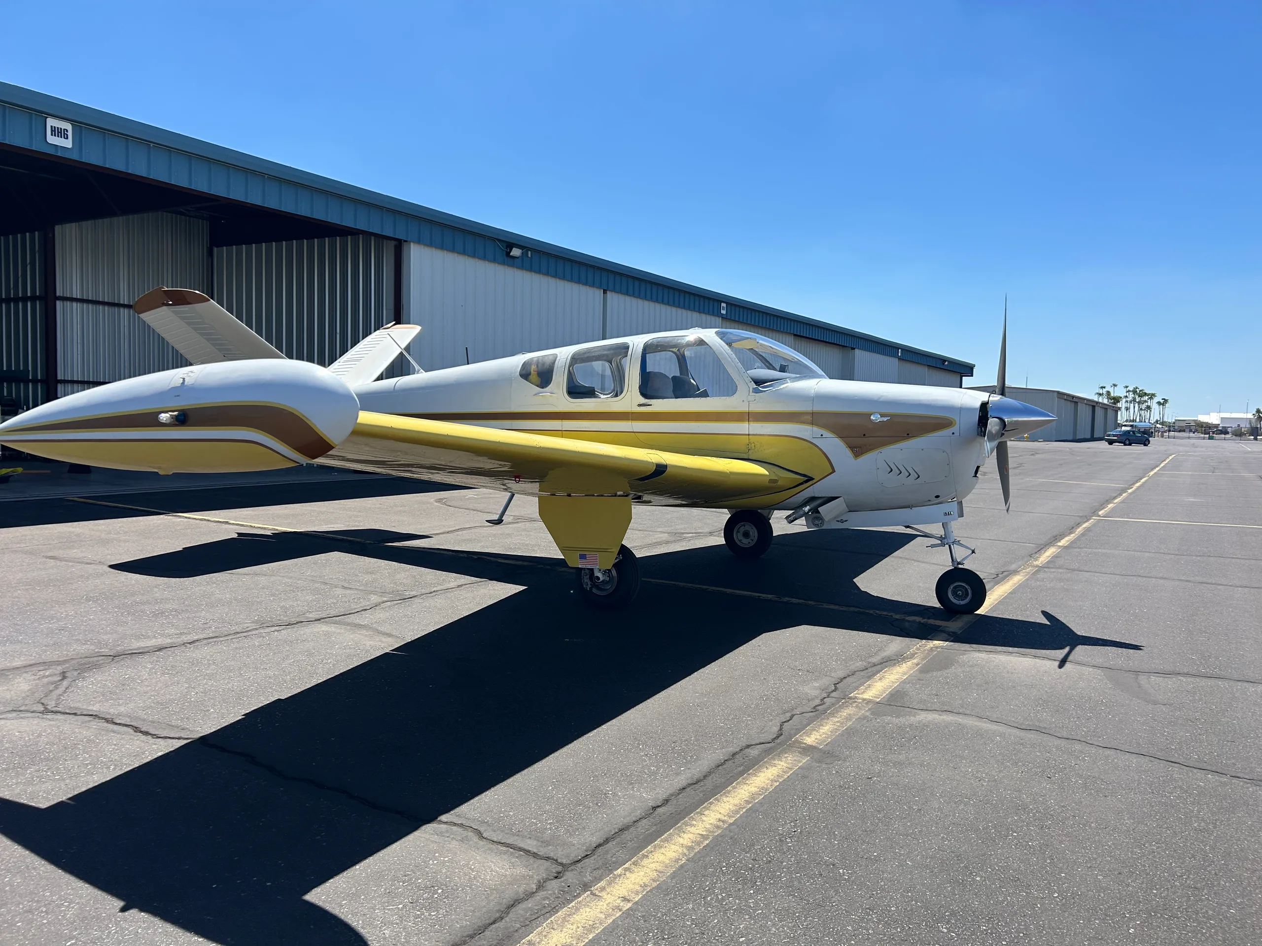 A white and yellow single-engine propeller aircraft with brown accents is parked on an asphalt tarmac next to a hangar under a clear blue sky.