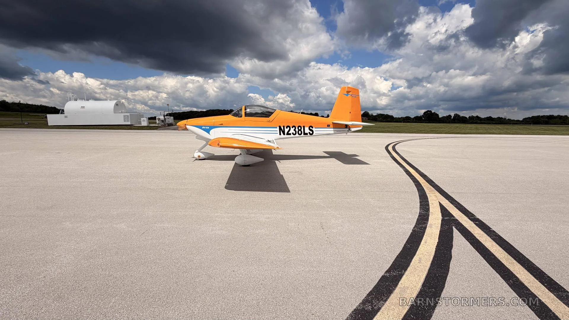 A side view of an orange and white single-engine aircraft with registration N238LS on an airfield, with dark clouds overhead.
