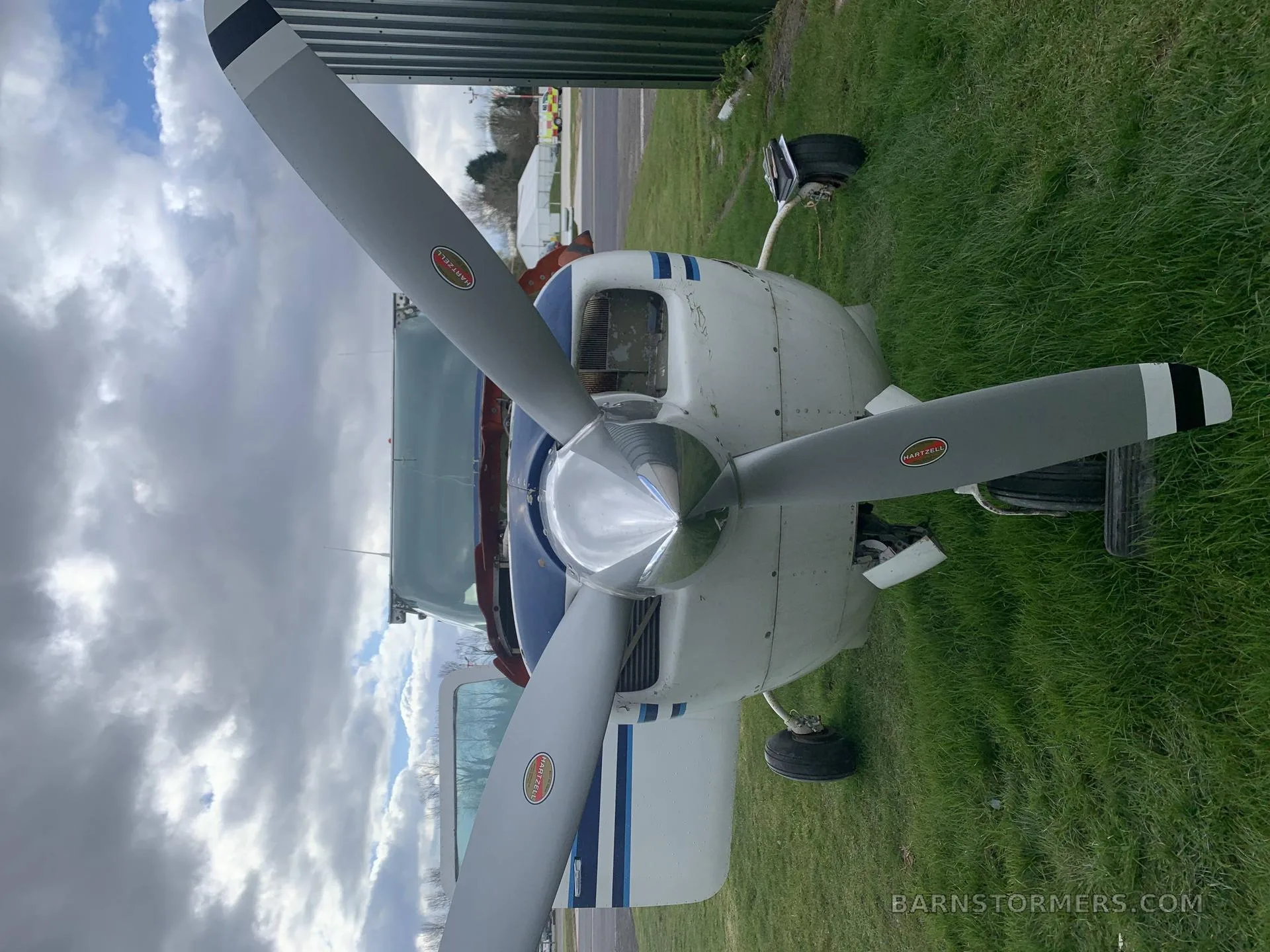 A front view of a white aircraft with blue stripes and a three-bladed propeller, parked on a grassy field under a cloudy sky.