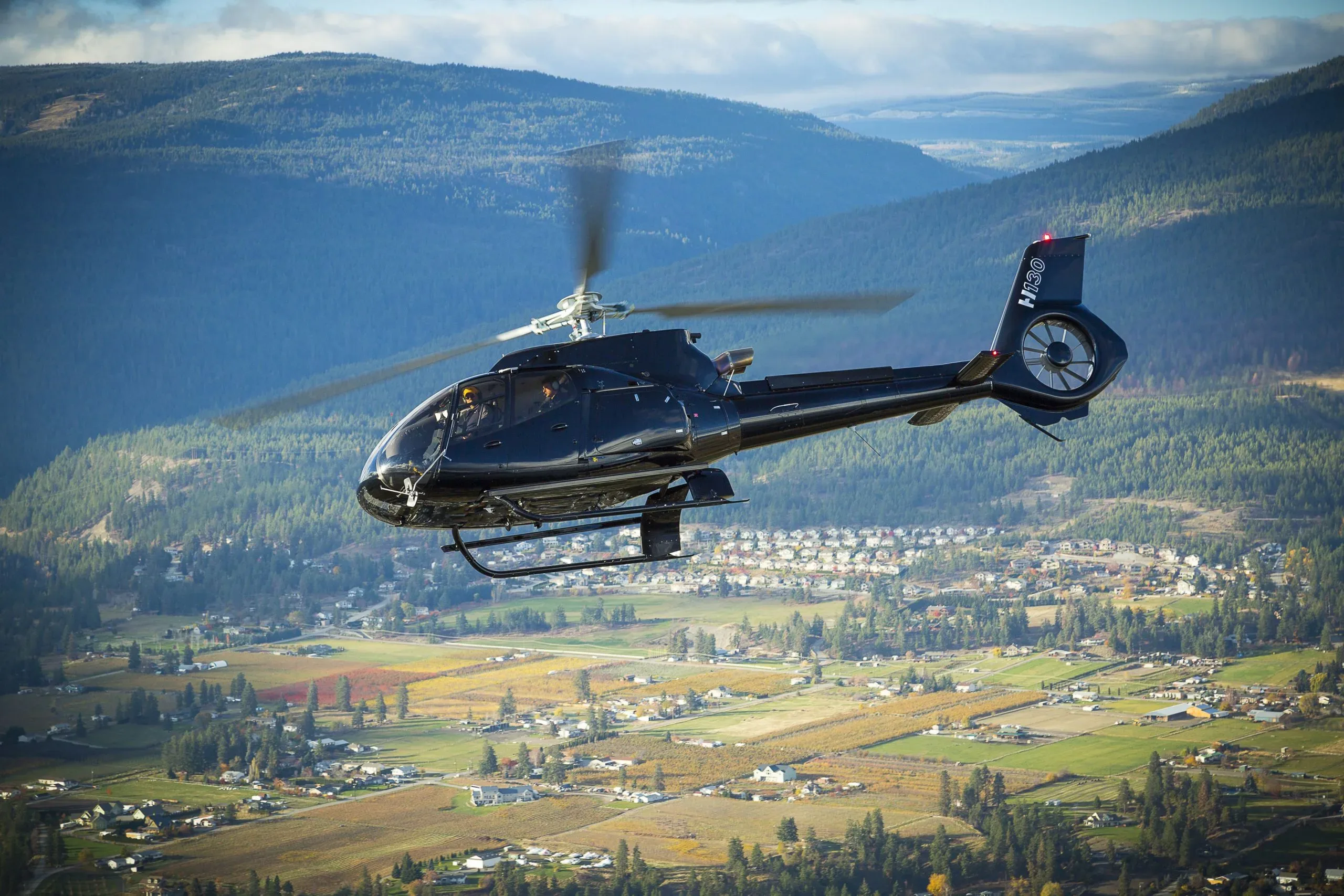 A black Airbus H130 helicopter with two pilots flying over a scenic valley with a town and forested mountains.