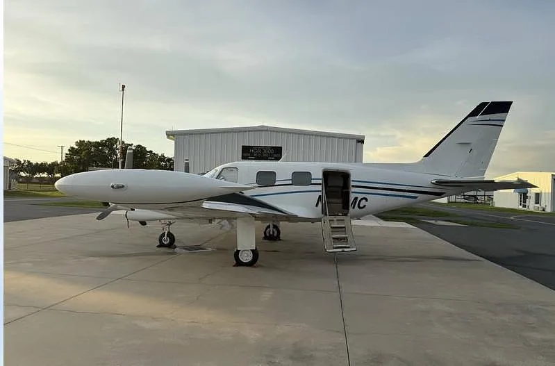 A white twin-engine propeller aircraft with blue and black accents, registration N53MC, parked on a tarmac with its passenger door open.