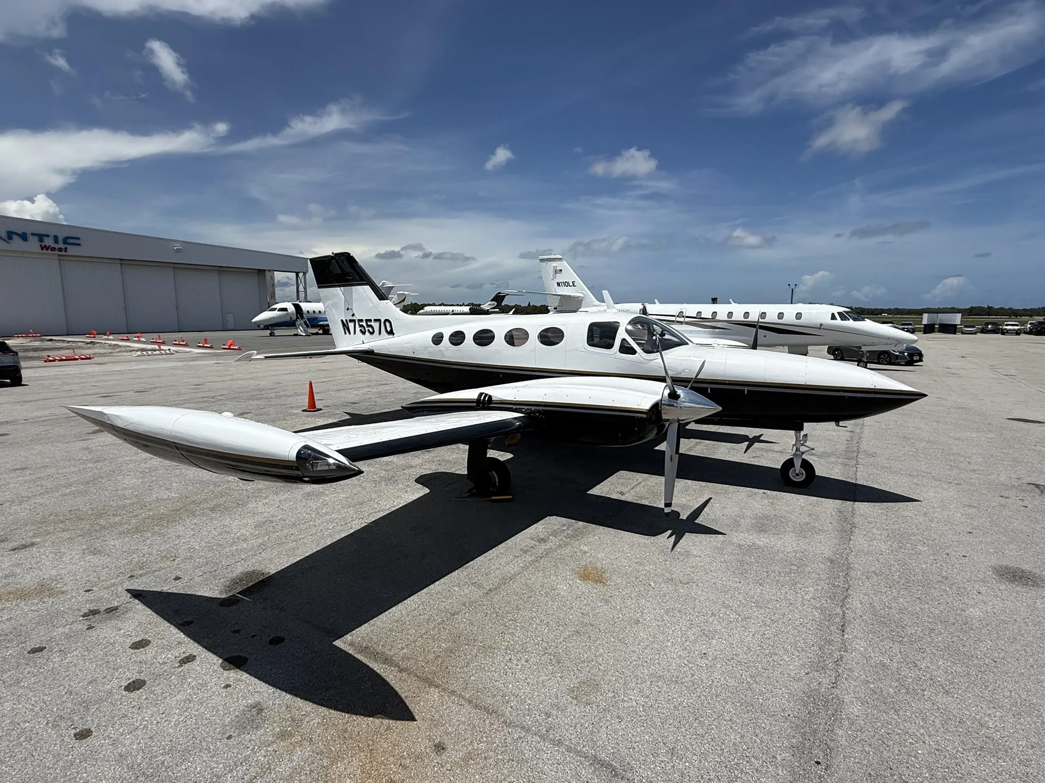A side view of the white and black twin-engine propeller aircraft N7557Q parked on an airfield with other aircraft.