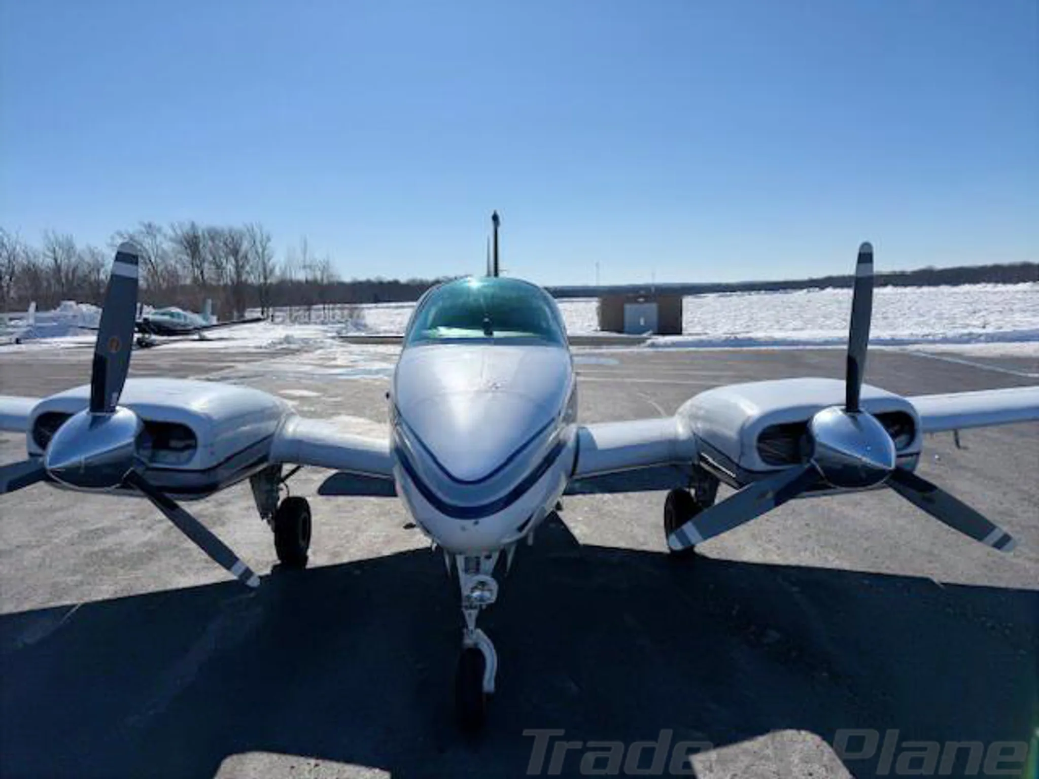 Head-on view of a white twin-engine aircraft with blue and grey stripes on a snowy tarmac.