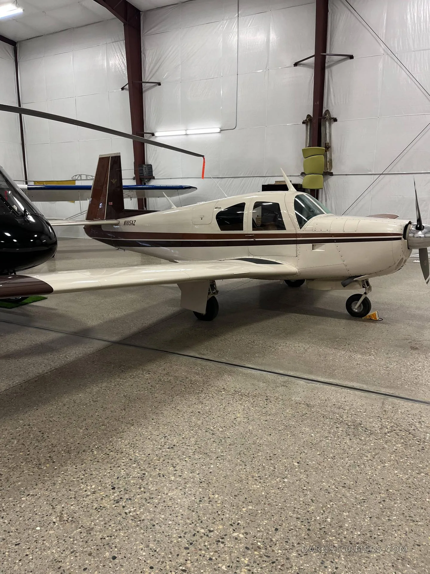 A cream and brown single-engine propeller aircraft (Beechcraft Debonair N165AZ) is parked in a hangar, with a helicopter partially visible on the left.