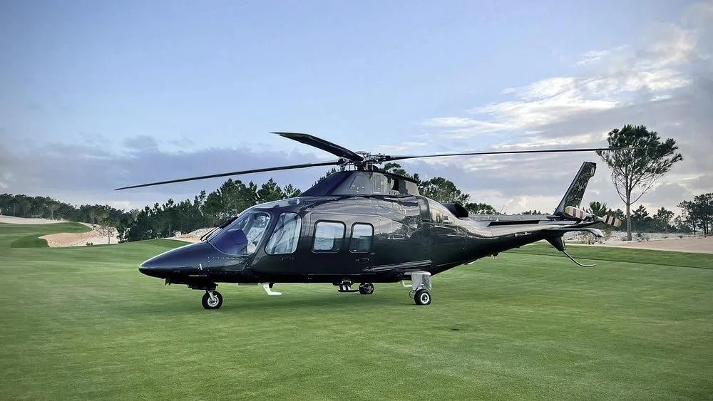 A dark grey helicopter with its main rotor blades extended, parked on a green golf course with trees and sand traps in the background under a partly cloudy sky.