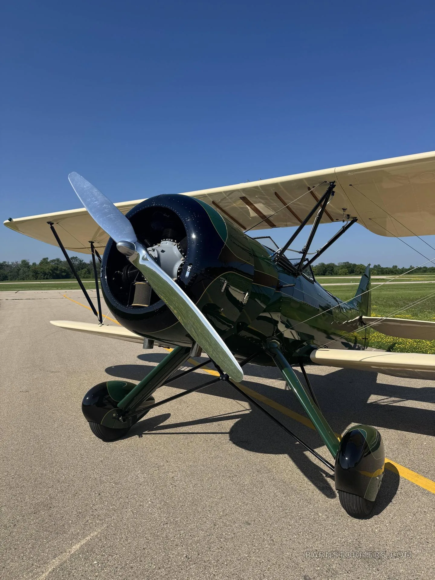A close-up front view of a green and cream biplane, highlighting its propeller and landing gear on an asphalt runway.