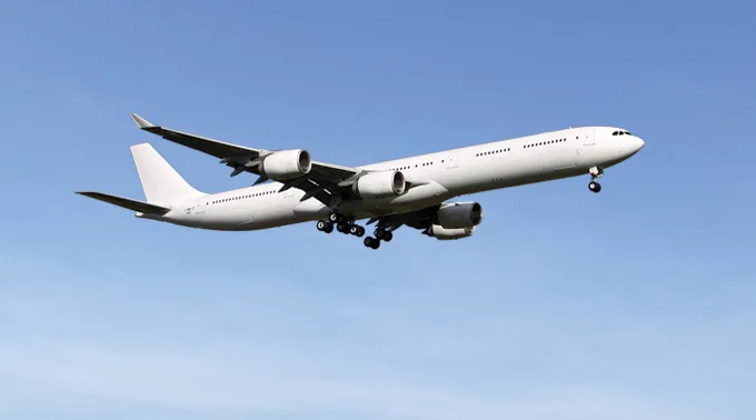 A large, white four-engine passenger jet with landing gear extended flies against a clear blue sky.