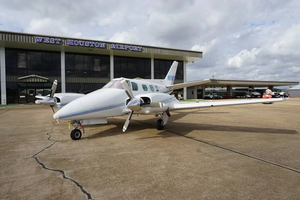 A white twin-engine propeller aircraft with blue stripes, registration N818KT, parked on the tarmac in front of the West Houston Airport building.