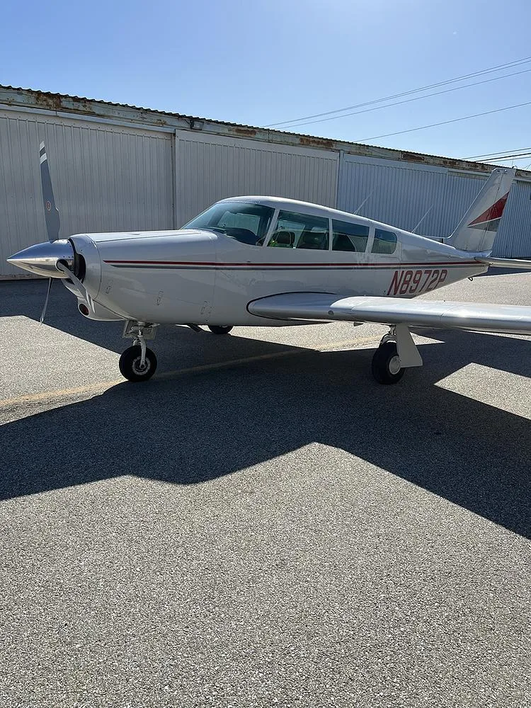 A three-quarter front view of the white Piper Cherokee 180 (N8972P) with red and grey stripes parked on an asphalt ramp.