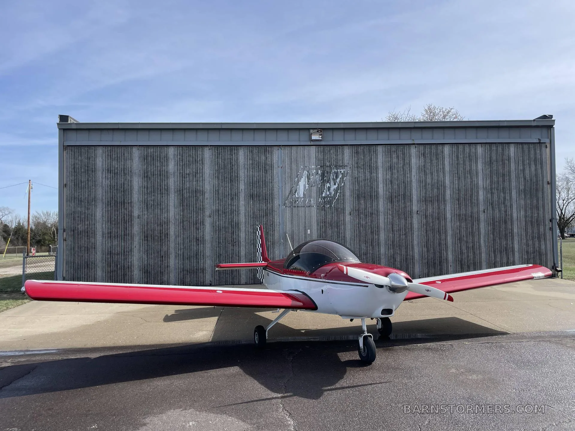 A red and white single-engine aircraft with a checkered tail is parked on an asphalt apron in front of a large metal hangar.