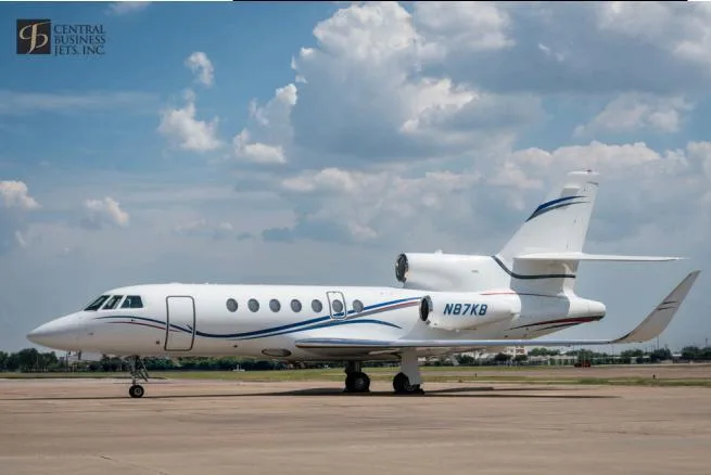 A white Dassault Falcon 900 business jet with blue and red stripes, registration N87KB, parked on an airport tarmac under a partly cloudy sky.