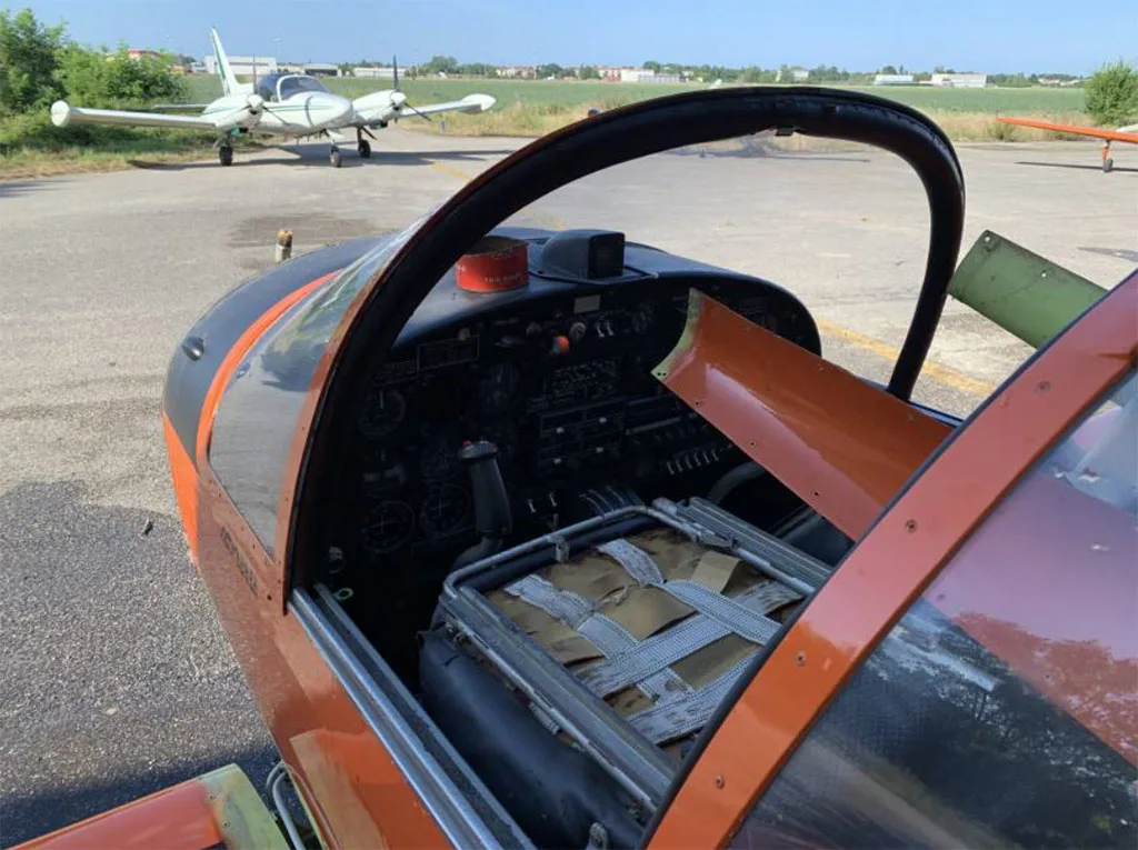 The open cockpit of an orange aircraft, showing the instrument panel and a strapped-down cargo area, with another aircraft in the background.