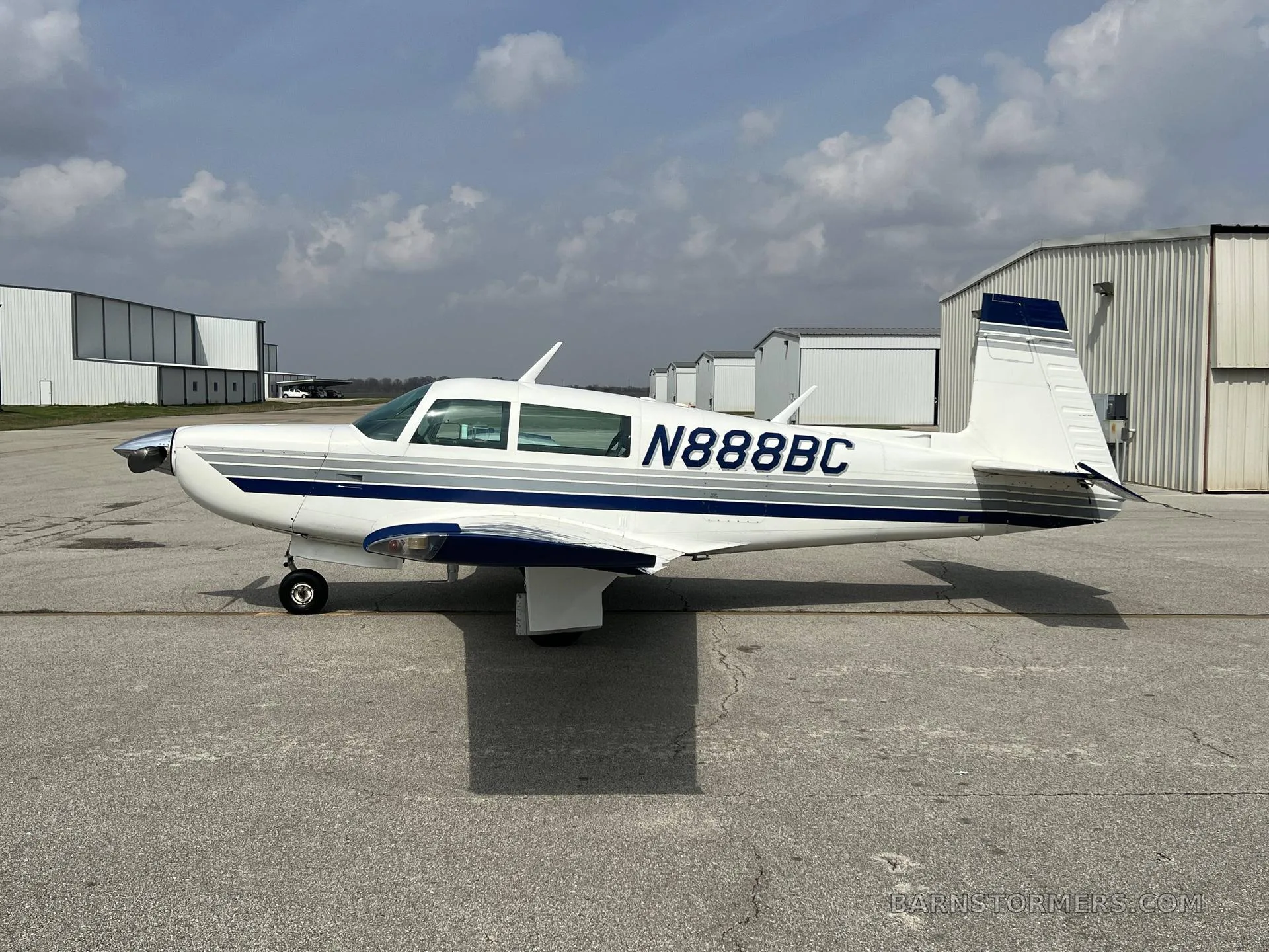 A white single-engine aircraft with blue and grey stripes (N888BC) parked on an asphalt ramp with hangars in the background.