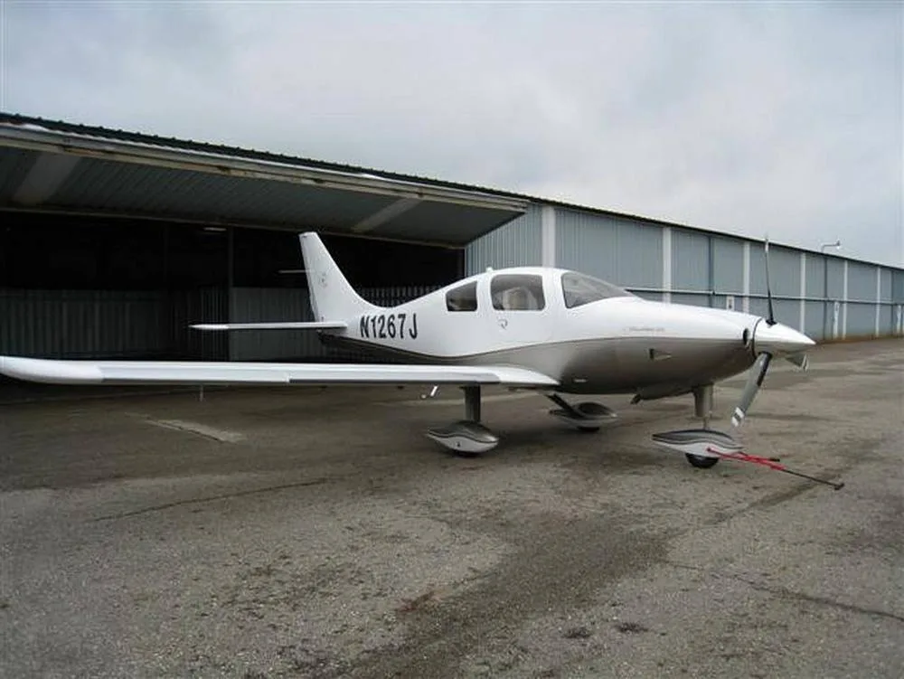 A white and silver Columbia 350 aircraft, N1267J, parked on an asphalt apron next to a hangar under an overcast sky.
